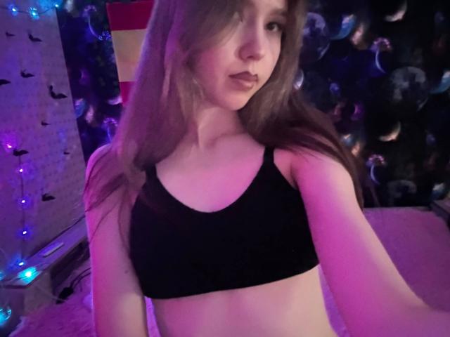 AprylVanochten with long brown hair, wearing a black sports bra, showing her small tits and slim body, in a purple-lit room with string lights.