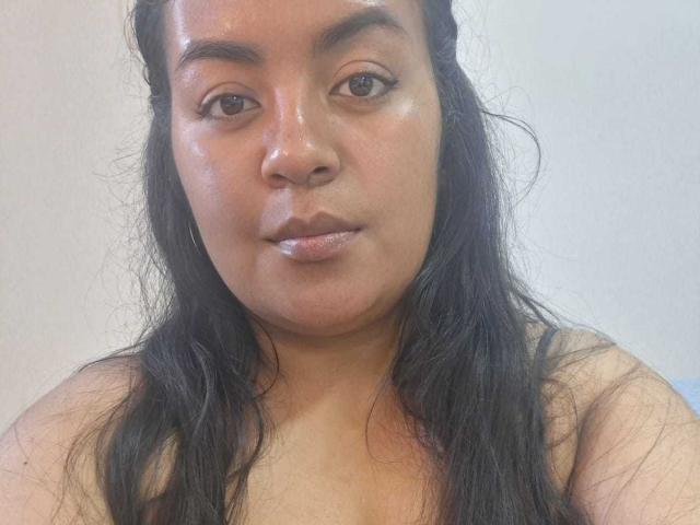 AthenaTheas has shiny brown skin, dark wet hair, and a neutral expression. She's topless with visible small, round tits and a soft, slightly chubby face.