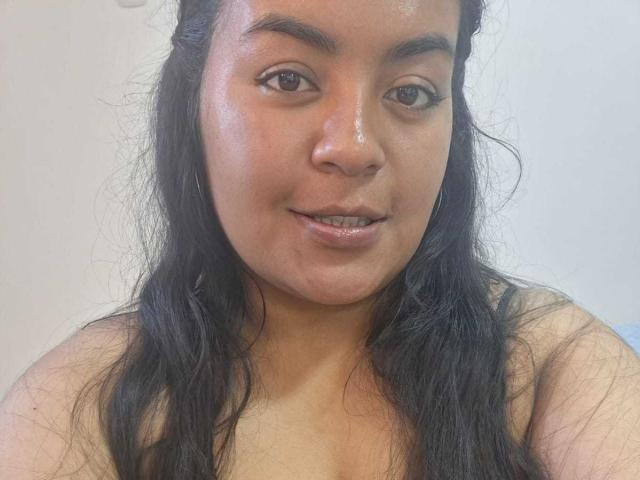 AthenaTheas has a chubby face with shiny skin, black wavy hair, brown eyes, and a slight smile, showing her upper body and naked shoulders.
