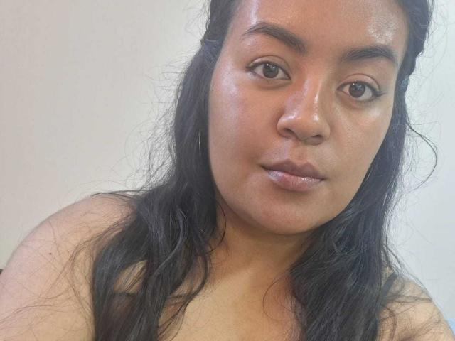 AthenaTheas has shiny, dark brown skin and long black hair, with a neutral expression and subtle, glossy lips. She has small, round brown eyes and is looking at the camera.