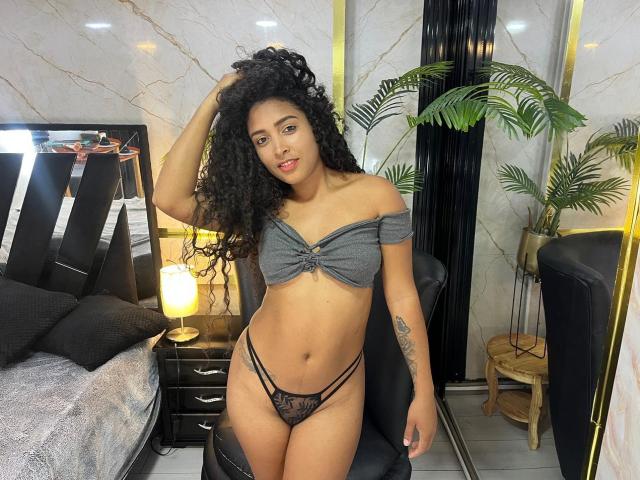 PaulinaRizzo with curly black hair, tan skin, small tits, wearing a gray off-shoulder top and black lace panties, standing in a bedroom with a black chair.