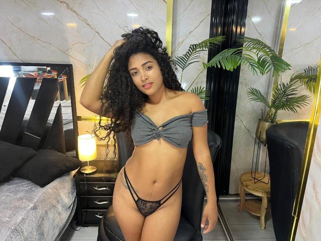 PaulinaRizzo with curly black hair, wearing a gray off-shoulder top and black lace thong, showing her tits and pussy, standing in a bedroom with a lamp and plants.