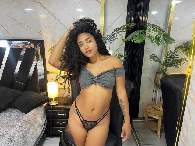 PaulinaRizzo with curly black hair, gray off-shoulder top, and black lace thong, standing in bedroom, hand in hair, tattoo on right arm, medium tits, brown skin.