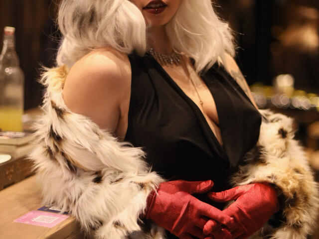 Mettis with white curly hair, deep black dress showing tits, red gloves, fur coat, dark lipstick, necklace, and a bottle in the blurry background.