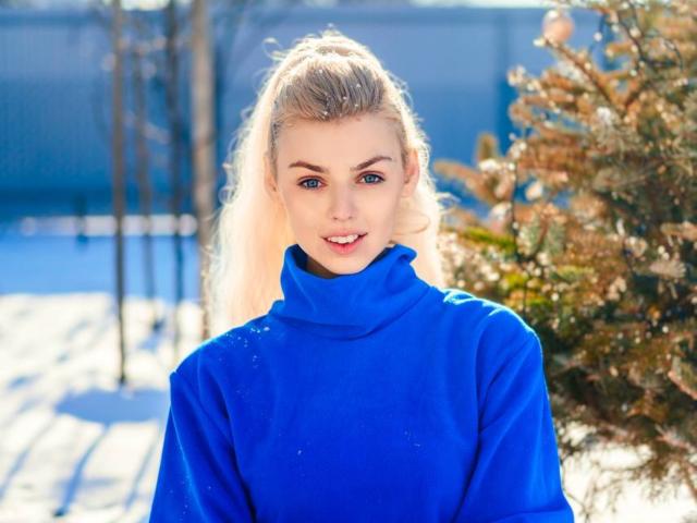 Sanda Hayslett, avec une queue de cheval blonde, des yeux bleus et un sourire éclatant, portant un pull bleu à col roulé, se tient dans la neige, avec un sapin à droite..