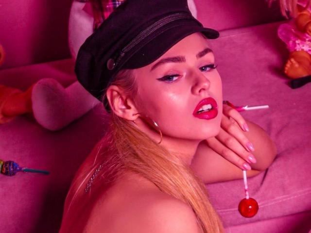 SandaHayslett met blond haar, een zwarte muts, rode lippenstift en oorringen, die een suikerstok vasthoudt, naakt, roze nagels, verleidelijke blik, roze verlichting, een roze bank, sexy, schattig..