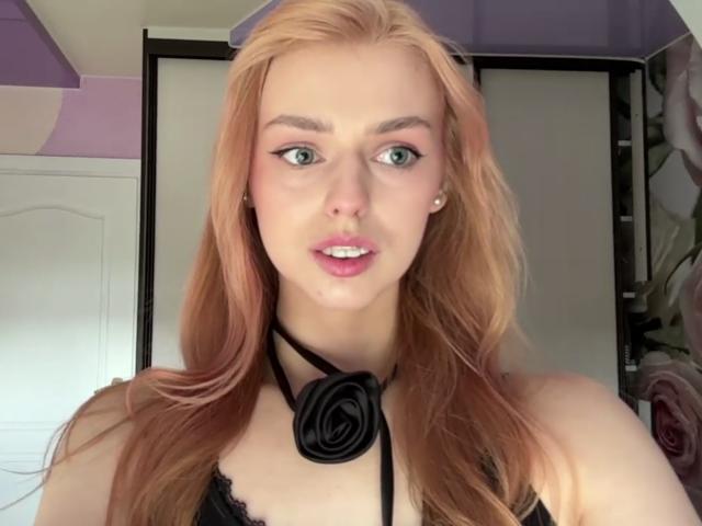 Sanda Hayslett, con lunghi capelli biondo-rosati, occhi verdi, labbra rosa, una collana nera con un ciondolo circolare, indossa una camicetta di pizzo nero che lascia intravedere leggermente il seno..