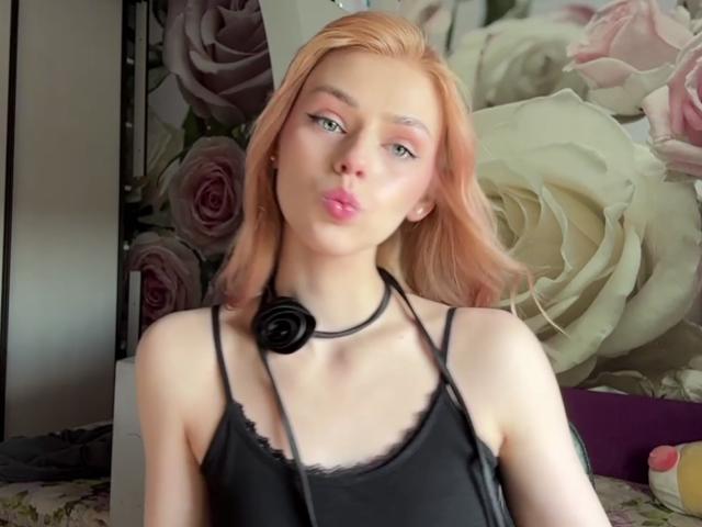 Sanda Hayslett, com cabelo rosa, blusa de alça preta, colar com rosa preta, lábios rosados curvados, pele clara, maquiagem suave, olhando para a câmera, fundo floral, rosas rosa e brancas..