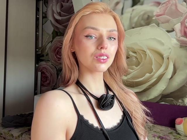 SandaHayslett hat blondes Haar, blaue Augen und rosafarbene Lippen. Sie trägt ein schwarzes Tanktop und ein schwarzes Headset, das um ihren Hals hängt, wobei ihre Brüste leicht zu sehen sind..