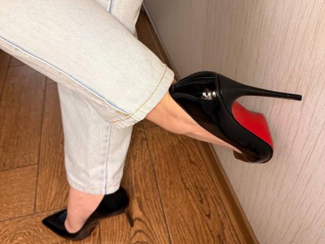 KaiaLein's foot in a black stiletto with red soles, wearing white jeans and black socks, standing on wooden floor, holding the heel.
