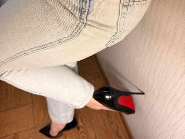 KaiaLein's ass in tight white jeans, wearing black high heels with red soles, standing on wooden floor, touching a white wall.