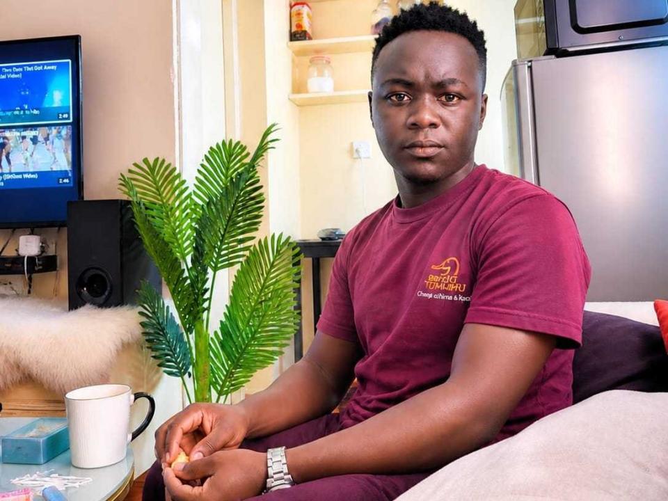 EbonyTjay, um homem de pele escura com cabelo preto curto, vestindo uma camiseta vermelha, sentado em um sofá, com uma expressão séria, segurando um pequeno lanche, uma caneca branca em uma mesa..