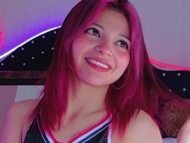 ShofiaWett with pink hair, smiling, wearing a blue and red cheerleader top, hoop earrings, looking up, pink and purple neon lights in background.