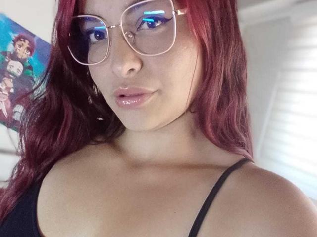 MarilynRouse with red hair, wearing glasses, black tank top, showing her tits, shiny pink lips, light skin, in a selfie. Her ass is partially visible.