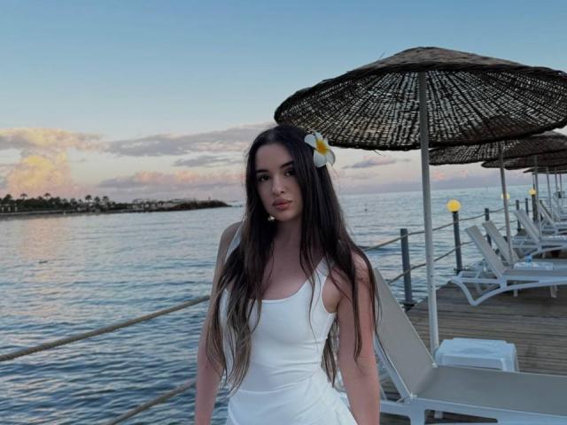 AllieViv with long dark hair, white dress, and plumeria in her hair, standing on a wooden dock, next to a woven umbrella, at sunset, with calm water.