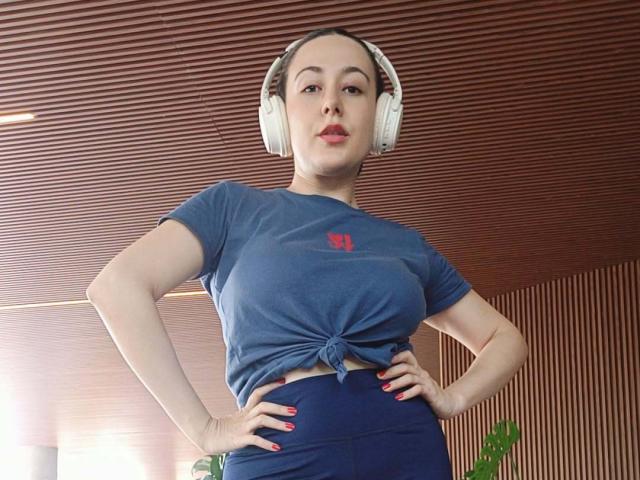 MabyLegrand with headphones, blue tied shirt, showing tits, hands on hips, red nails, confident, looking down, wood ceiling, medium skin, brown hair, lipstick.