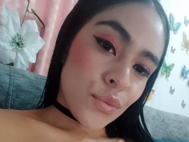 StellaBeckker has pink eye makeup, black choker, nose ring, and glossy lips, with black hair and a slight smile, showing her pouty tits in a selfie.