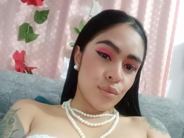 StellaBeckker with pink eyeshadow, pearl necklace, and stud earrings, black hair, nose ring, tattoos on shoulders, looking at camera, sitting on gray couch.