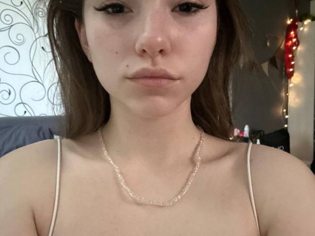 TericaSarley has pale skin, brown eyes, and long brown hair. She's wearing a thin white spaghetti strap top and a delicate necklace. Her small tits are visible.