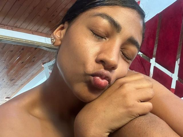 NatalyBlonder with closed eyes, pouting lips, and brown skin, resting her chin on her knee, wearing a gold hoop earring, under a wooden ceiling with a red umbrella.
