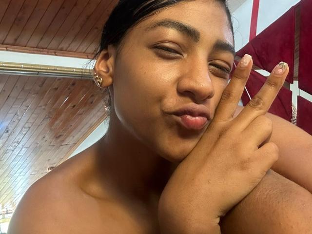 NatalyBlonder, brown-skinned, making a peace sign near her face, has small nose, full lips, and black hair, wearing gold earrings, in a wood-ceilinged room, looking cute and confident.