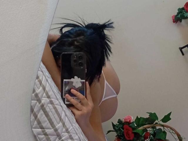 KatheLux with black hair in a messy bun, taking a mirror selfie with her phone, showing her big tits in a white bra, holding a quilted mattress.