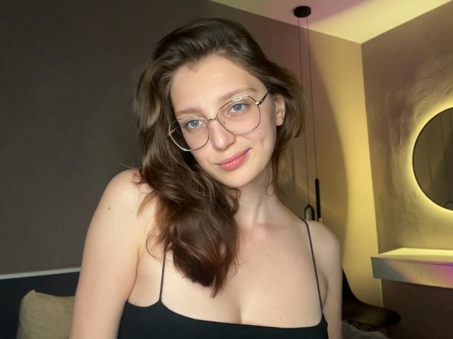 StaceyAverbach with wavy brown hair, glasses, black spaghetti strap top, showing small tits, light skin, slight smile, and blue eyes, in dimly lit room.