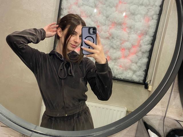 Heminuel with dark brown hair in a black hoodie and skirt, holding a phone, smiling at a mirror, showing a cute ass, in a selfie.