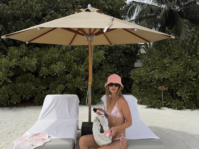 TheHottestBlonde, wearing pink bikini and pink hat, sits under beach umbrella with white loungers, holding a pink towel, has small tits, and is holding a black bag.
