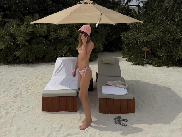 TheHottestBlonde stands on white sand, wearing pink beanie, white bikini, showing her small tits and slim ass, under beige umbrella, with gray lounge chair and white towel.