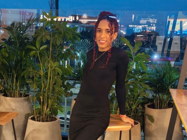 TiffannyDolly in a black dress, standing with one arm on a wooden table, smiling, with red curly hair, and a necklace, surrounded by potted plants at dusk.