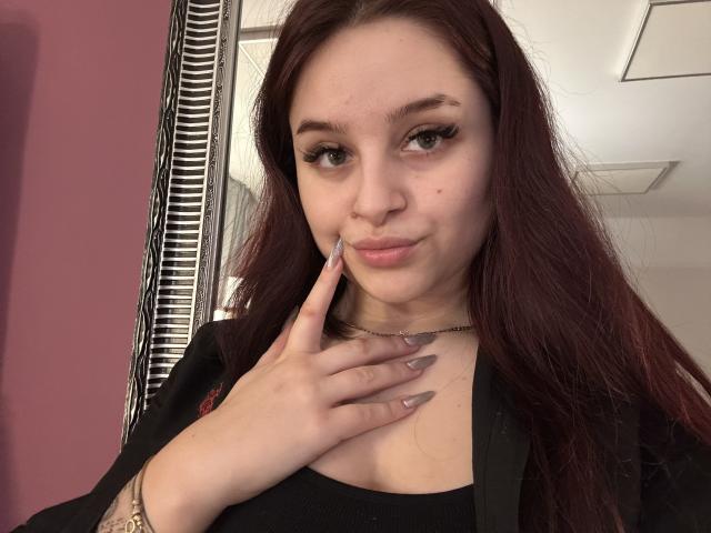 Lucie, mit langen roten Haaren, trägt ein schwarzes Oberteil und hält ihren Finger in der Nähe ihrer vollen Lippen, wobei sie ihre langen, grauen Nägel und ein goldenes Armband zur Schau stellt..