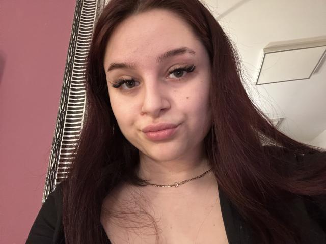 LucieRed with long red hair, subtle makeup, and a small black mole on her left cheek, wearing a black top and a thin necklace, slightly smiling.