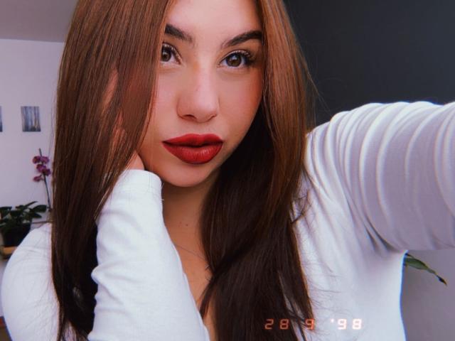 VenusxMusa has long brown hair, red lipstick, and is wearing a white long-sleeve top, with her hand near her face in a selfie. Her brown eyes are focused on camera.