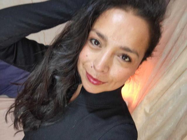 StangelarRivera with dark wavy hair, smiling, wearing a black turtleneck, nose ring, and red lipstick, lying on a bed, head slightly tilted, looking at camera.
