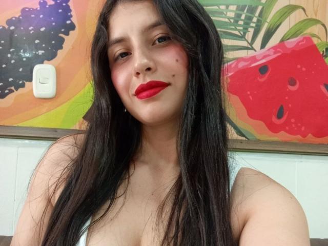 MadissonFontaine has long black hair, red lipstick, and a cute face with a small mole on her left cheek, wearing a white top, showing her tits slightly.
