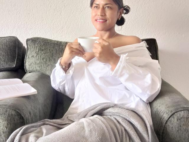 IvannaDuran with bare shoulders, white shirt off, holding a white cup, gray blanket, and book on green couch, smiling, dark hair in bun, casual, relaxed, cozy.