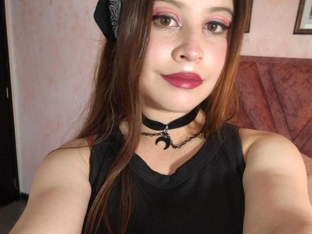 MelaniBecker has brown eyes, pink eyeshadow, red lipstick, black choker with crescent moon, black tank top, and brown hair, looking at the camera with a slight smile.