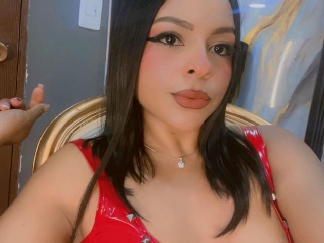 LindaFrancoo with dark brown hair, red latex top, thick black eyeliner, pink blush, and orange lipstick, holding a hand near her face. Her boobs are visible.