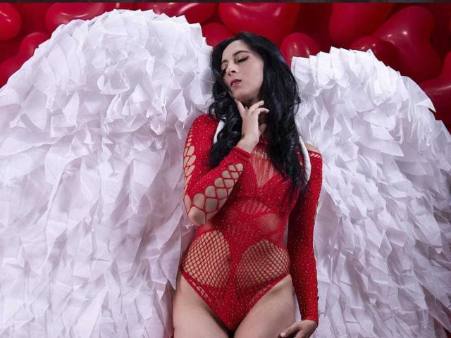 AlessiaRinaldii in red, fishnet, winged bodysuit, with large white, textured wings, black wavy hair, and a finger touching her chin, showing her tits and ass.