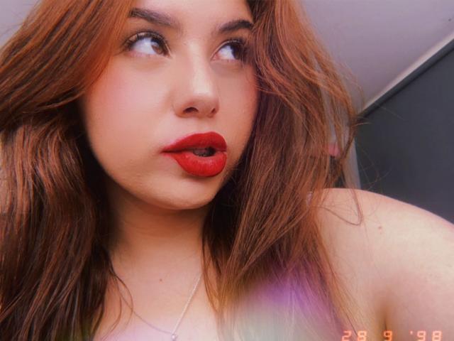 VenusxMusa heeft rode lippenstift, volle rode lippen en een neusring. Ze kijkt opzij met bruine ogen en heeft lang, golvend, roodbruin haar. Haar borsten zijn klein..