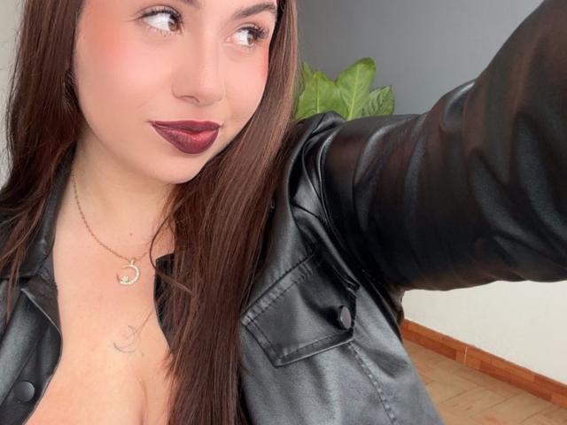 VenusxMusa with long brown hair, dark lipstick, black leather jacket, necklace, and cleavage, taking a selfie, showing a small tattoo on her chest.