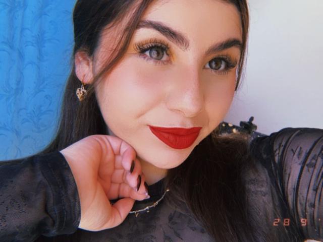 VenusxMusa with red lipstick, black sheer top, long brown hair, green eyes, and heart-shaped earrings, holding her chin, smiling. Her tits are not visible. Date "28 9" in corner.