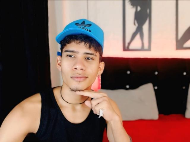 SkinnyCuteAlyer with short curly black hair, wearing a blue Adidas cap, black tank top, and choker, touching chin with finger, pink ear gauges, and black sleeve ring.