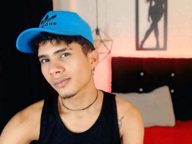 SkinnyCuteAlyer, tan-skinned, short black hair, blue Adidas cap, black tank top, thin black necklace, small nose, subtle smile, visible tattoo on right shoulder, sitting, red bed in background.