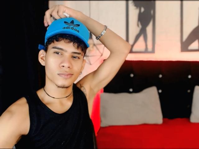 SkinnyCuteAlyer in a blue Adidas cap, black tank top, and choker, raising arm, showing armpit, bracelet on wrist, short curly black hair, and a slight smile.