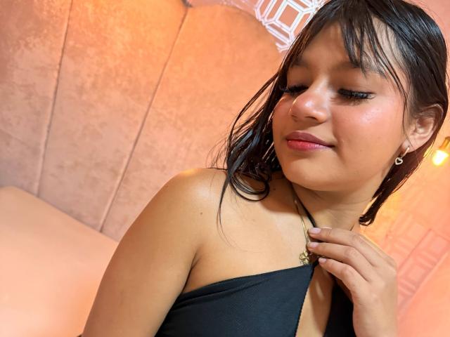 SamanthaLove with wet black hair, small round tits, wearing a black halter top, holding necklace, pink lipstick, subtle smile, pink ear studs, warm light, soft pink wall.