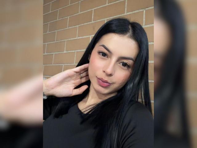 IsabellaJames has long black hair, brown eyes, and pink lipstick, wearing a black top, with a hand touching her cheek, reflecting in a mirror.