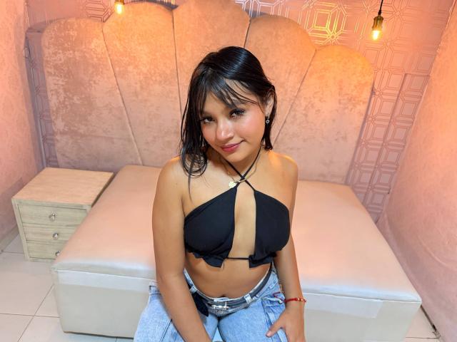 SamanthhaLove with wet black hair, small tits, wearing a black strappy top and grey pants, sitting on a white bed, smiling, light brown skin, red bracelet, and a necklace.