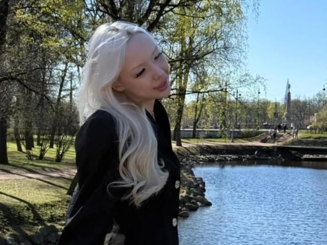 MargretMurak with long, platinum blonde hair, wearing a black button-up shirt, stands by a blue waterway, looking down with a slight smile. She's in a sunny park.